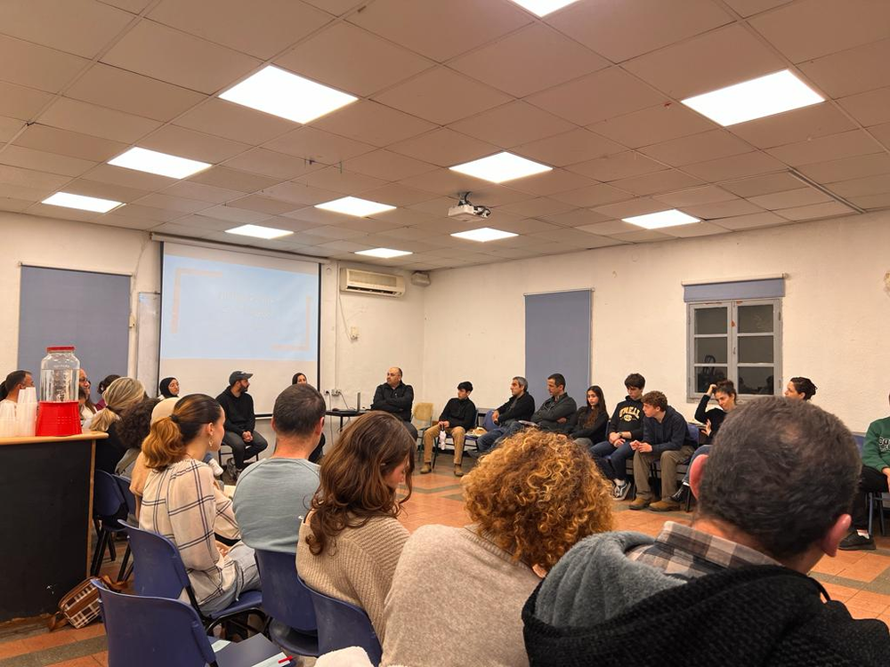 Around 25 teens and adults sit in chairs in a circle chatting