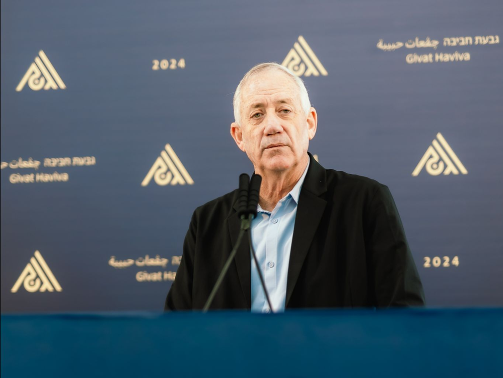 Benny Gantz standing behind a mic at a podium with givat haviva logos on the blue wall behind him