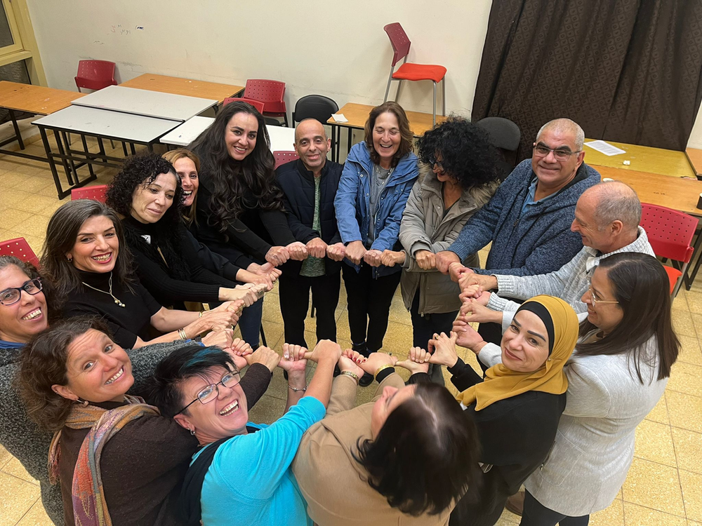 Teachers from the Shared Language program standing in a circle and smiling at the camera as they hold hands in the middle of a circle during a group activity