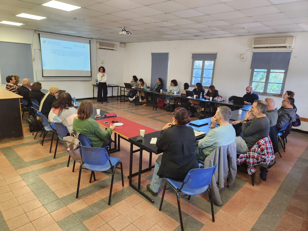 20 adults seated in chairs at tables in a U formation facing a wall with a slideshow projected onto it