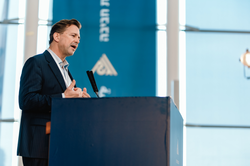 Steffen Seibert standing behind a mic at a podium with givat haviva logos on the blue wall behind him