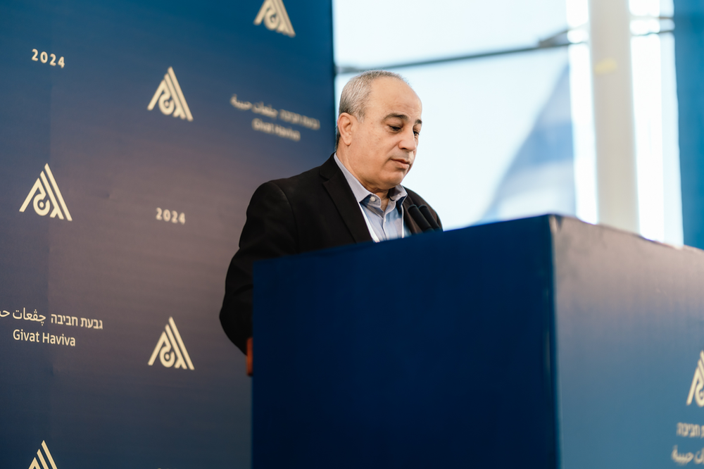 Mohammad Darawshe standing behind a mic at a podium with givat haviva logos on the blue wall behind him