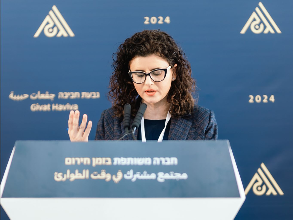 Dr. Nasreen Haddad Haj-Yahya standing behind a podium with a mic, givat haviva's logo is on the wall behind her