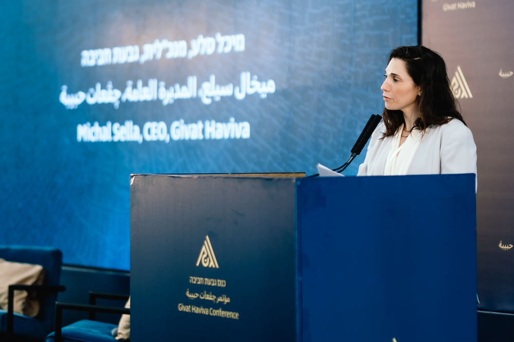 Michal Sella standing behind a mic at a podium with givat haviva logos on the blue wall behind him