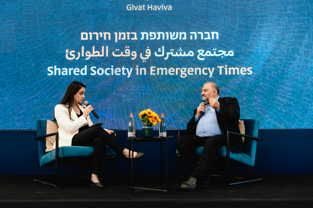 Mansour Avbas speaking with lior Kinnan. Both are stitting with microphones in their hands and the wall behind them has the title of the conference and Givat Haviva's logo