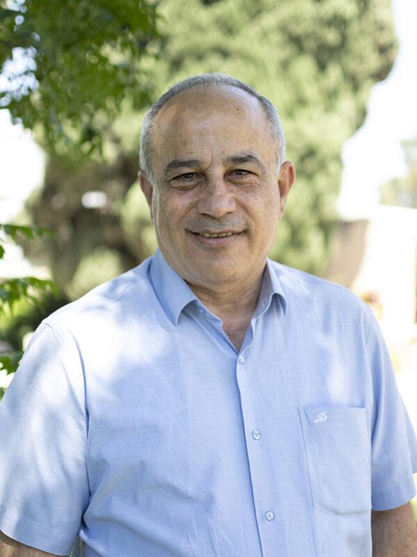 Mohammad Darawshe is standing outside and smiling at the camera. there are leaves behind him and he is in a light blue checkered shirt