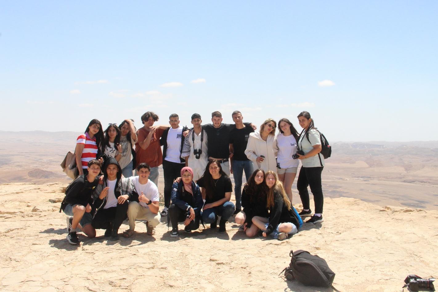 A group of students are standing at the edge of a viewpoint over a desert. some of them are holding cameras